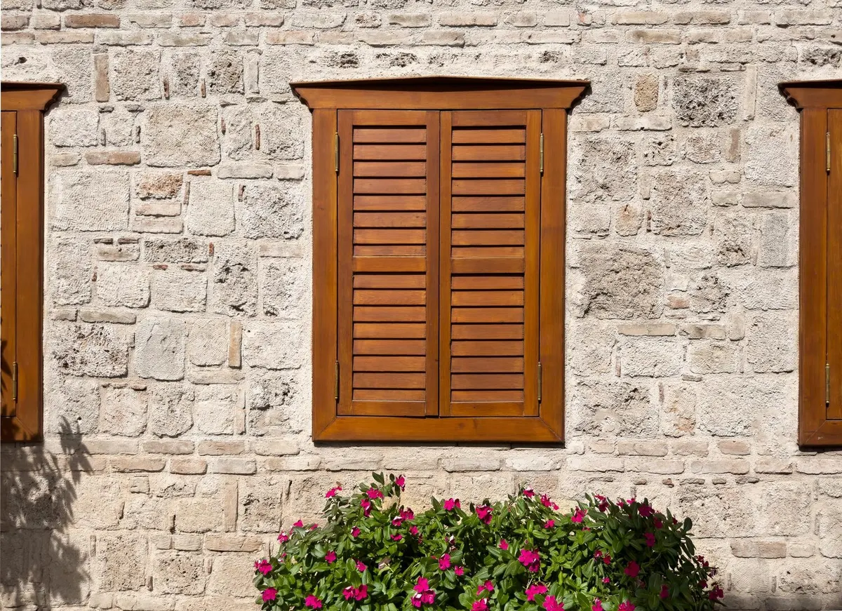 Volets à persiennes en bois sur un mur en pierres apparentes, jardinière fleurie au pied de la façade